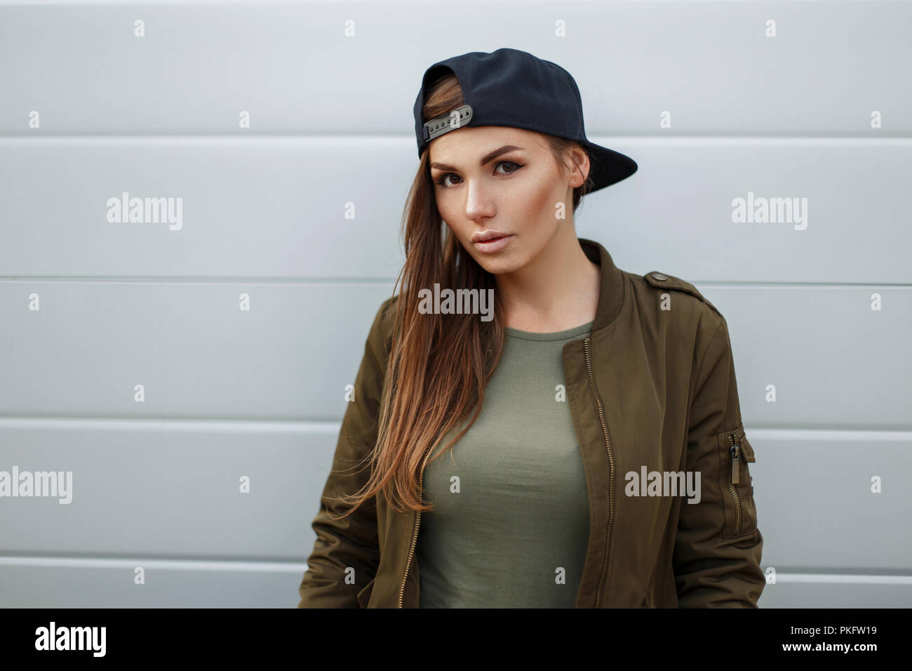 Calle retrato de una bella mujer con pecas en una gorra de béisbol en una chaqueta militar cerca de una pared metálica Foto de stock