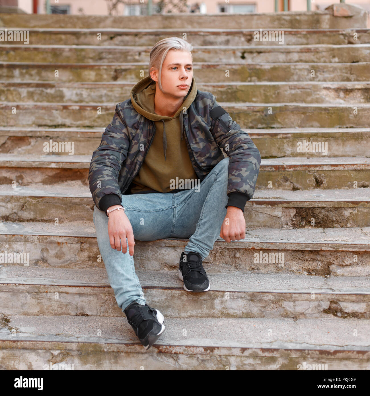 Moda hombre guapo con el pelo en una chaqueta militar con jeans y  zapatillas sentada en las escaleras Fotografía de stock - Alamy