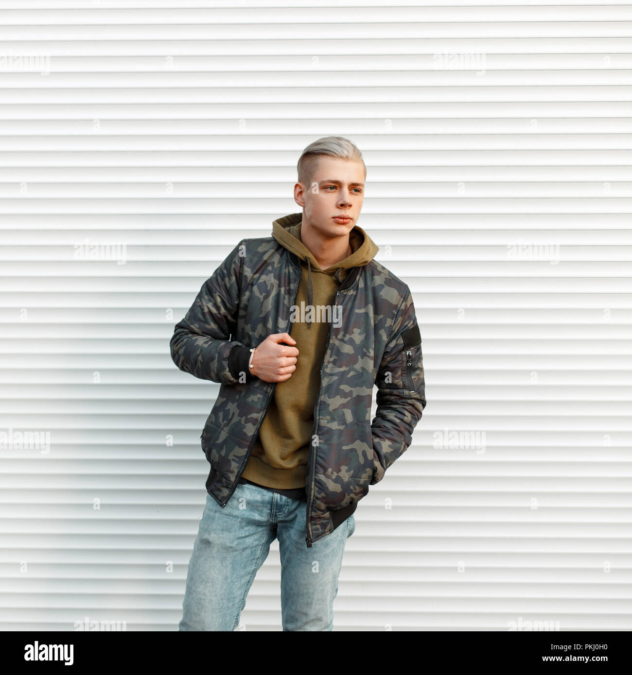 Guapo de moda moda hombre chaqueta militar con hoodie posando cerca de  pared metálica blanca Fotografía de stock - Alamy