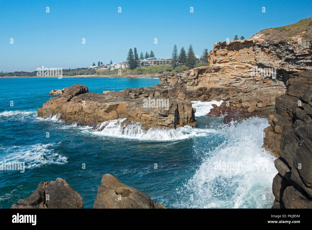 Las olas del Océano Pacífico rompiendo sobre las rocas al pie de los escarpados acantilados en Yamba con la ciudad en la distancia bajo un cielo azul, NSW, Australia Foto de stock