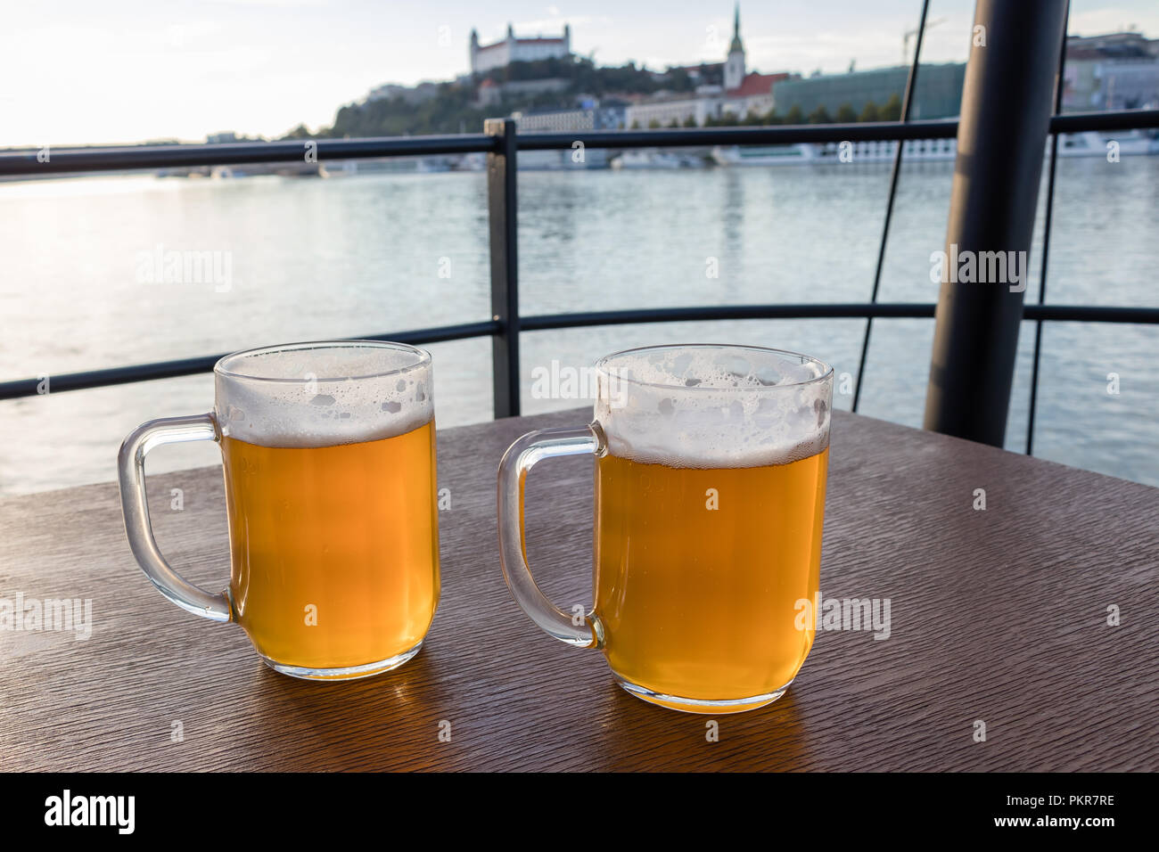 Jarras de cerveza colocados sobre una mesa de madera de fondo sobre el río Danubio y el castillo de Bratislava, Eslovaquia Foto de stock