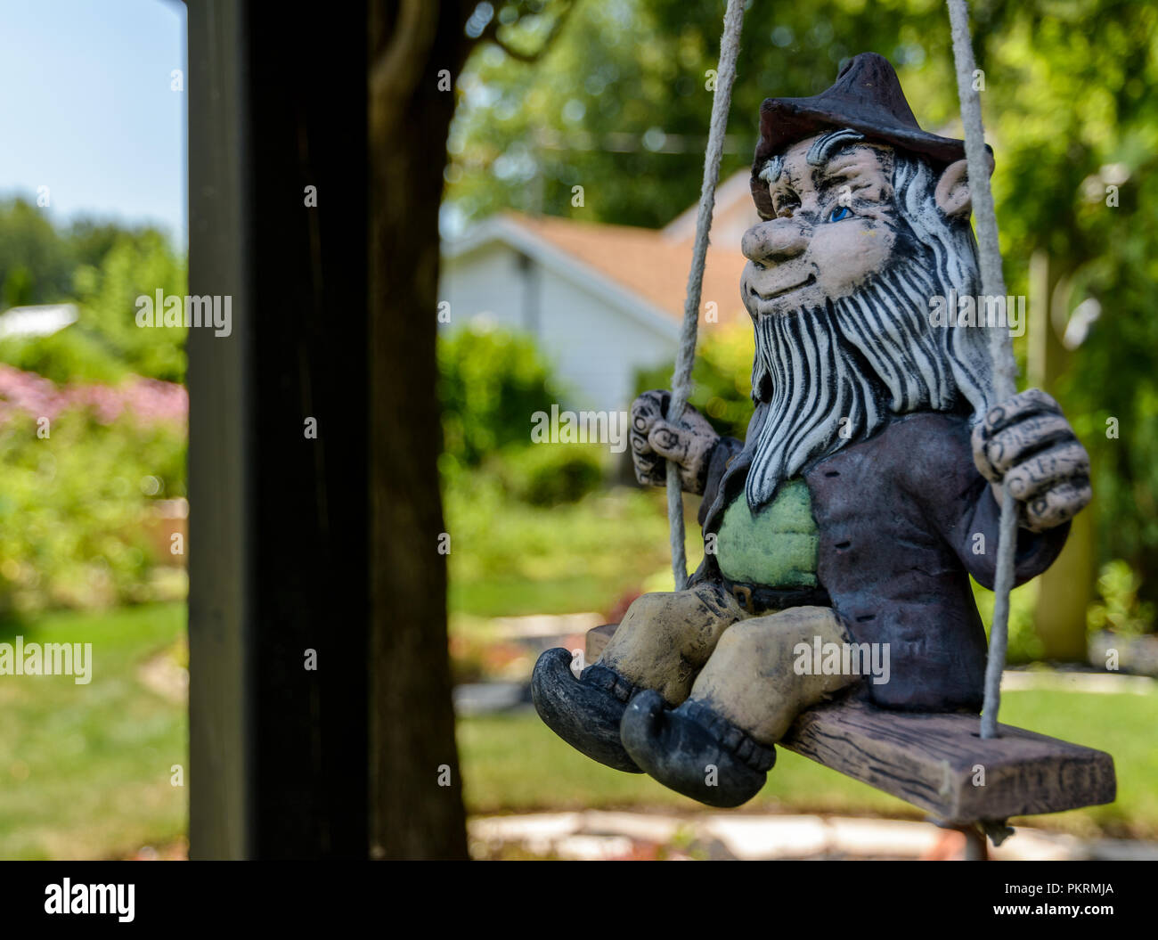 Macho de aspecto rústico Gnome con una barba blanca y un sombrero en un columpio con jardines en el fondo Foto de stock