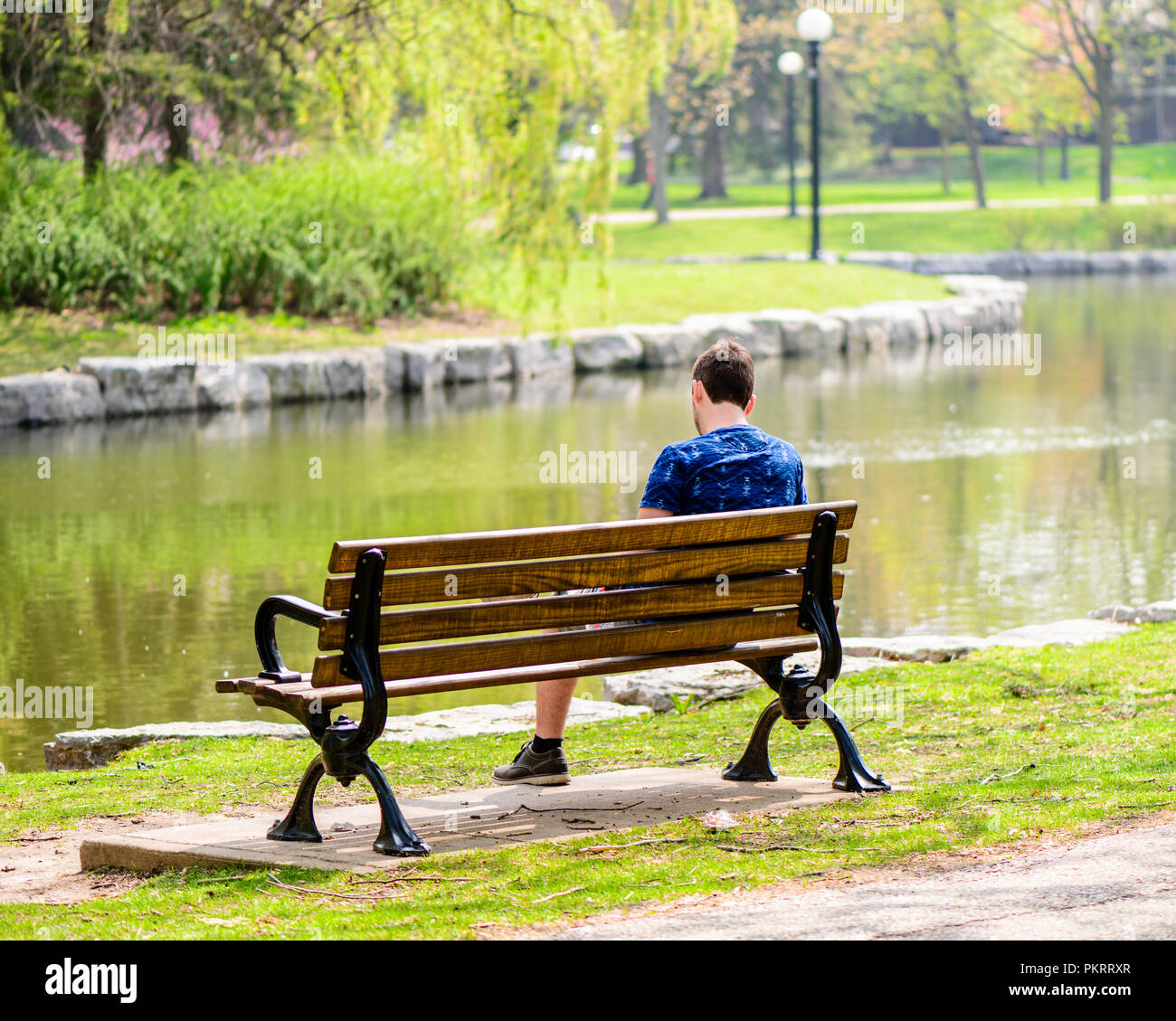 Hombre sentado en una banca del parque cerca de un río con árboles en el fondo Foto de stock