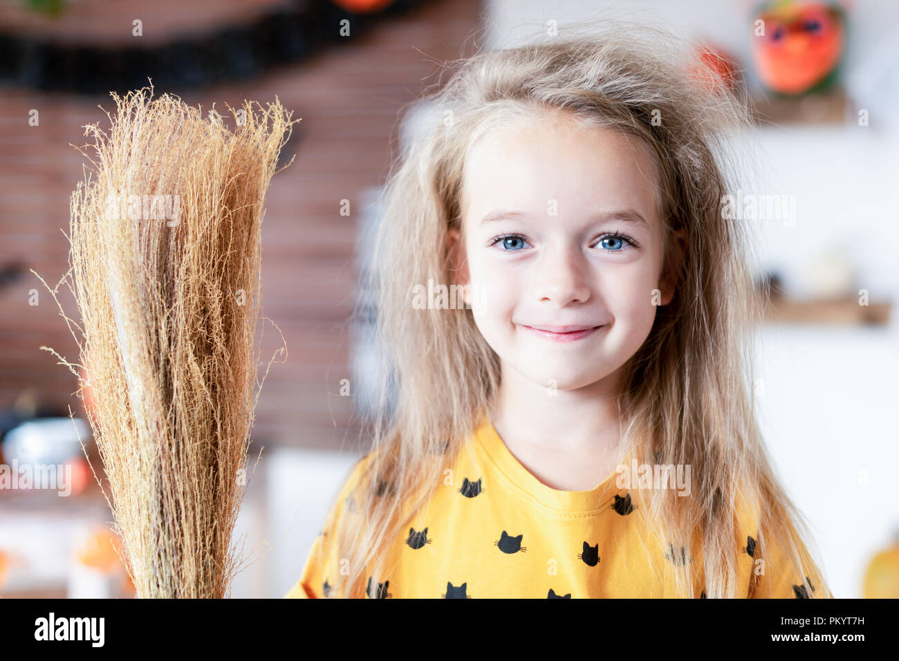 Cute Little Girl, con el cabello desordenado, vestida como una bruja y sosteniendo una escoba de pie en Halloween decorado salón, mirando a la cámara sonriendo. Foto de stock