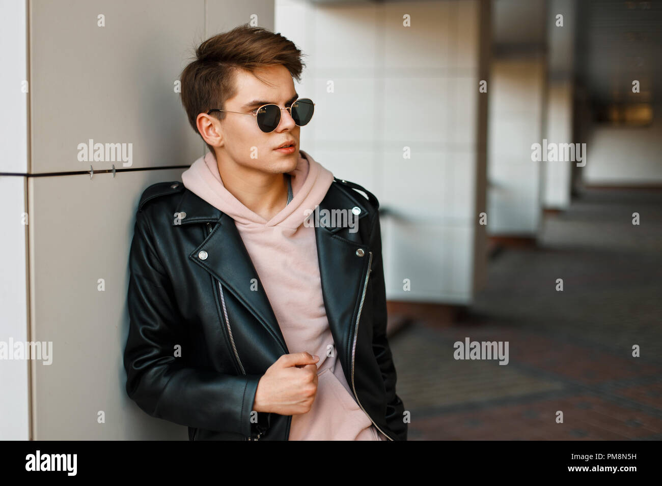 Joven apuesto hombre de aviador gafas de sol en una chaqueta de cuero negro  y una sudadera rosa cerca de una pared blanca moderna Fotografía de stock -  Alamy