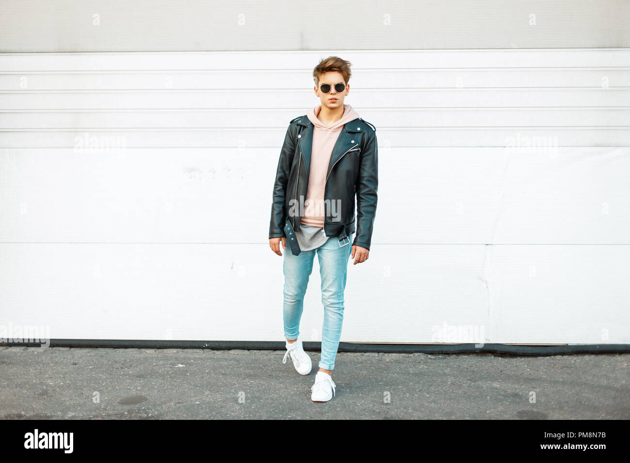 Un joven apuesto y elegante con gafas de sol en una chaqueta de cuero  negro, azul y blanco jeans de moda trendy sneakers cerca de una pared  metálica blanca Fotografía de stock -