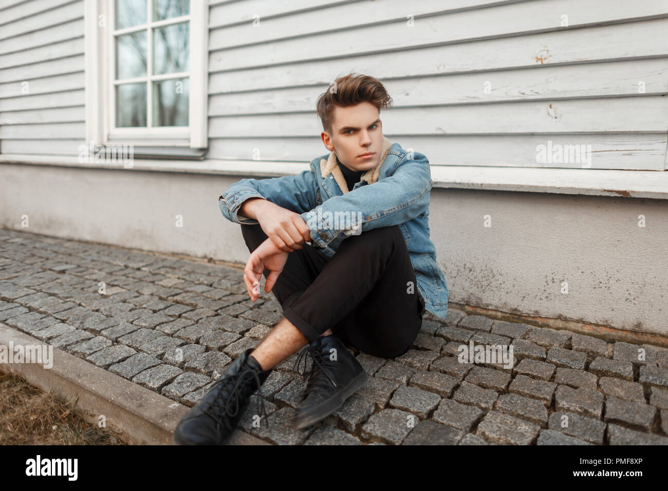 Modelo de moda joven hombre en elegante negro jeans y zapatillas sentada  cerca de una pared de madera Fotografía de stock - Alamy
