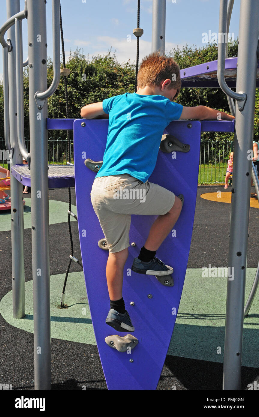 Siete-año-viejo muchacho escalar una pared de escalada hasta una diapositiva en un parque de juegos. Foto de stock