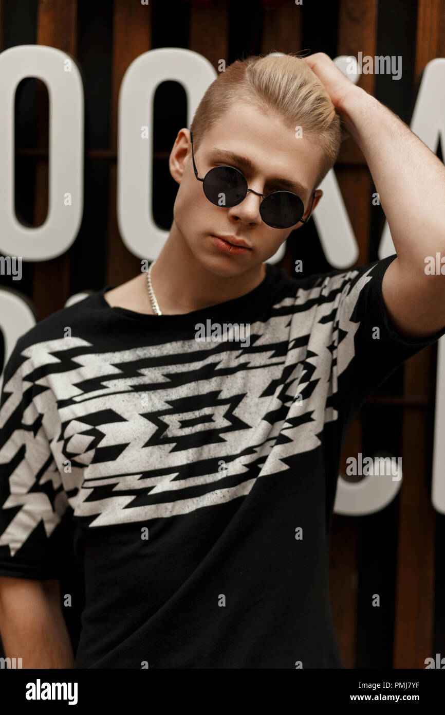 Elegante modelo joven hombre con un peinado en la ronda vintage gafas de  sol en una moda negro camiseta con soportes de impresión cerca de una pared  de madera con letras Fotografía