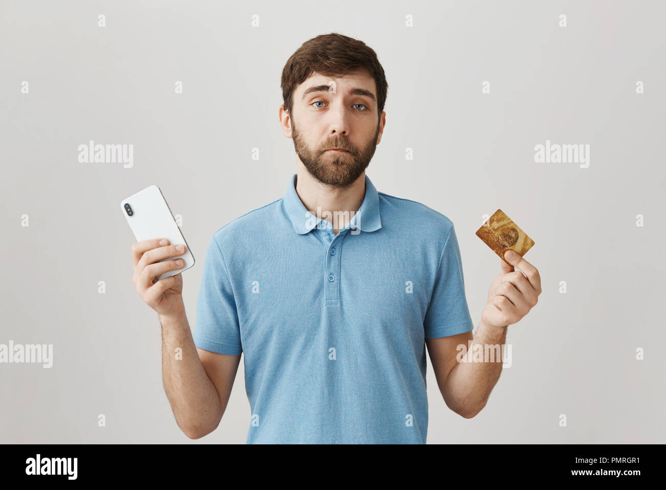 Inútil tarjeta de crédito con saldo insuficiente. Sombrío retrato del joven barbudo disgustó guy celebración smartphone y tarjeta bancaria, teniendo problemas con el pago en línea, erguido sobre fondo gris Foto de stock