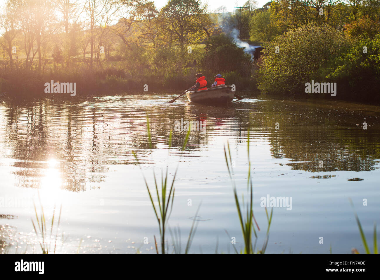 Dos niños remando un barco en un pequeño lago de vuelta iluminado por el sol de la noche. Foto de stock