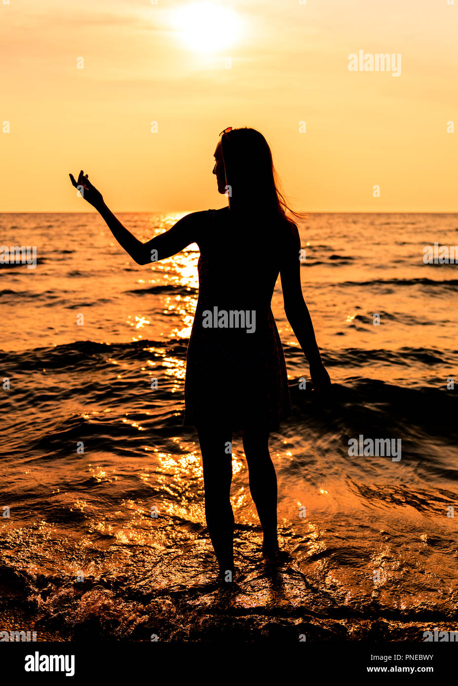 Jovencita en un vestido con pelo largo en la playa en silueta durante la puesta de sol con su mano extendida hacia el lado Foto de stock