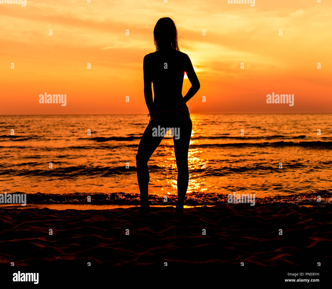 Jovencita en traje de baño con el pelo largo en la playa en silueta durante la puesta de sol en una pose casual Foto de stock