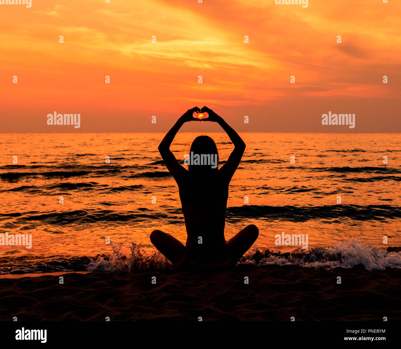 Jovencita en traje de baño con el pelo largo en la playa en silueta durante la puesta de sol haciendo un corazón con sus manos Foto de stock