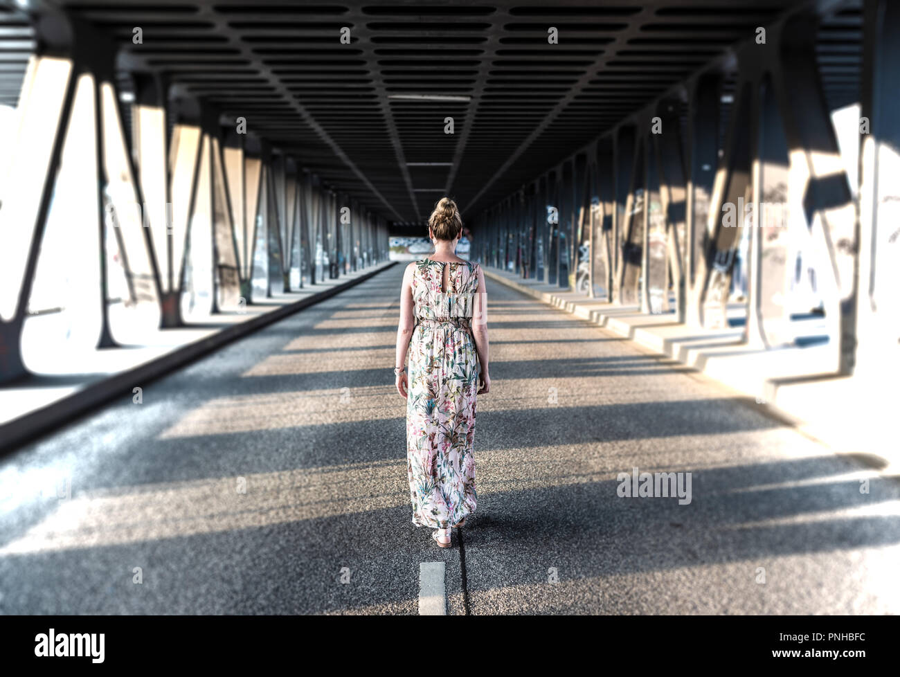 Vista trasera de una mujer en vestido largo caminar en medio de una carretera debajo del puente Foto de stock