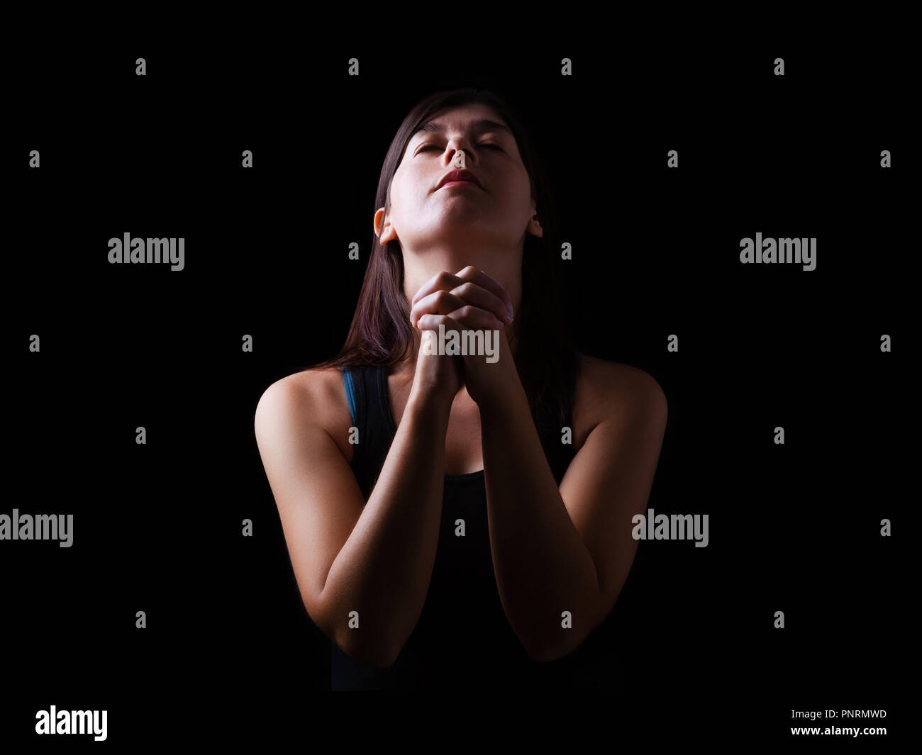 Mujer fiel orando, manos plegadas en el culto a Dios con la cabeza hacia arriba y los ojos cerrados de fervor religioso, sobre un fondo negro. El concepto de religión, Foto de stock