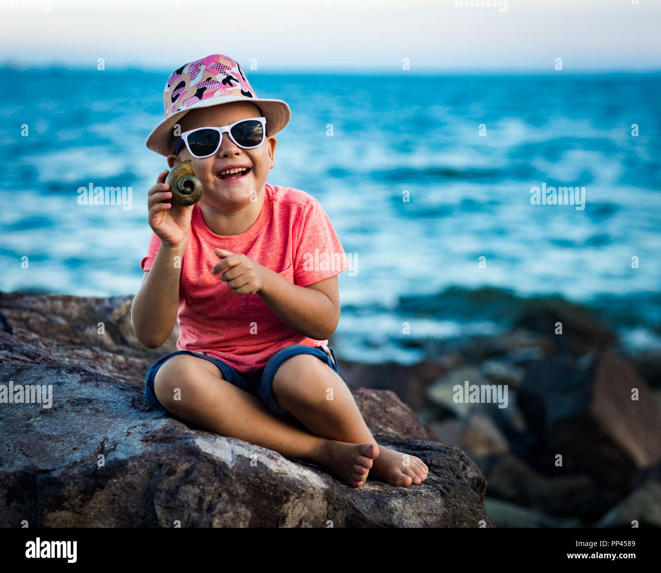 Chico con concha sentada sobre una roca cerca del mar Foto de stock