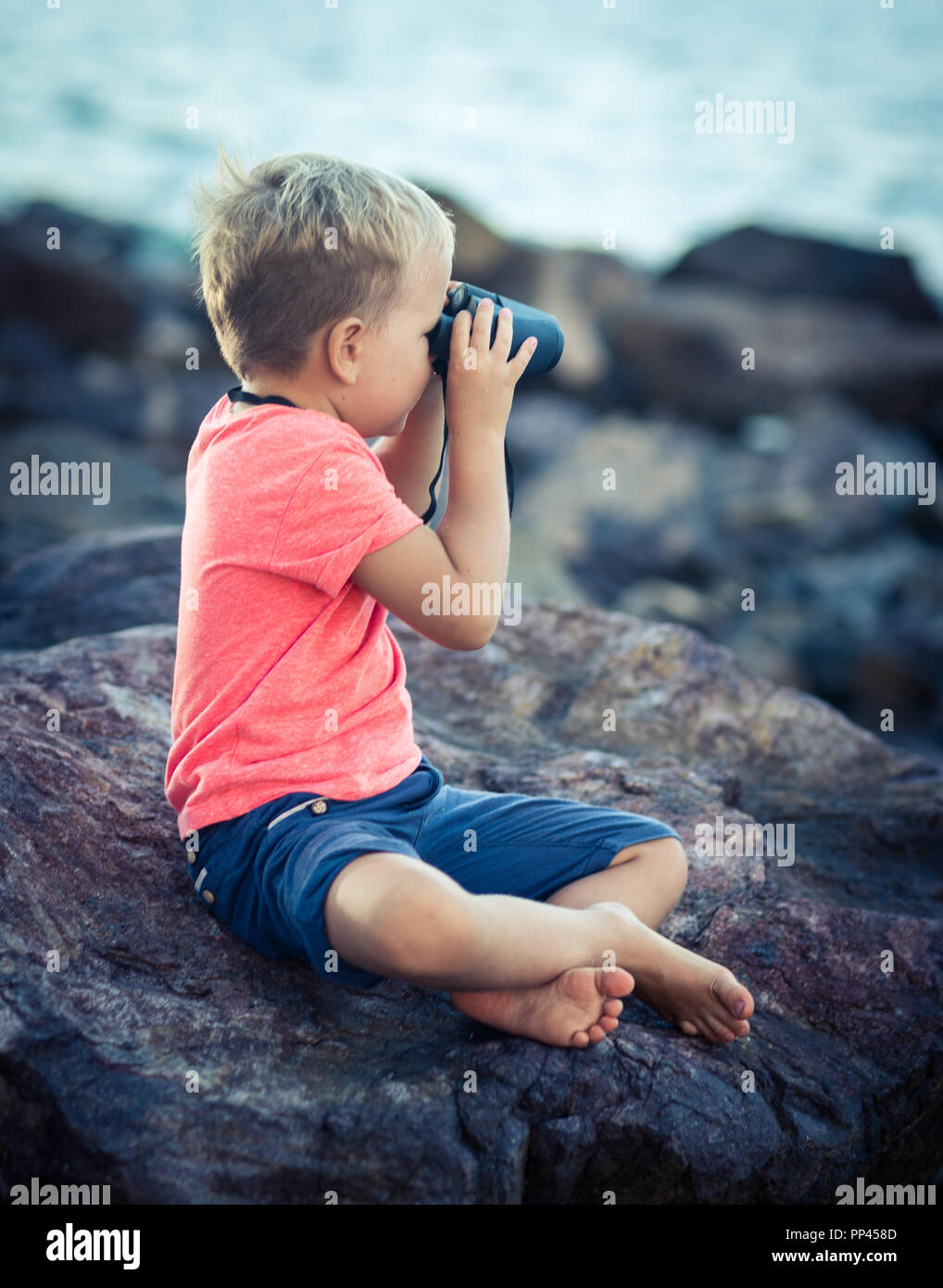 Chico busca lejos con binoculares, sentado sobre una roca cerca del mar Foto de stock
