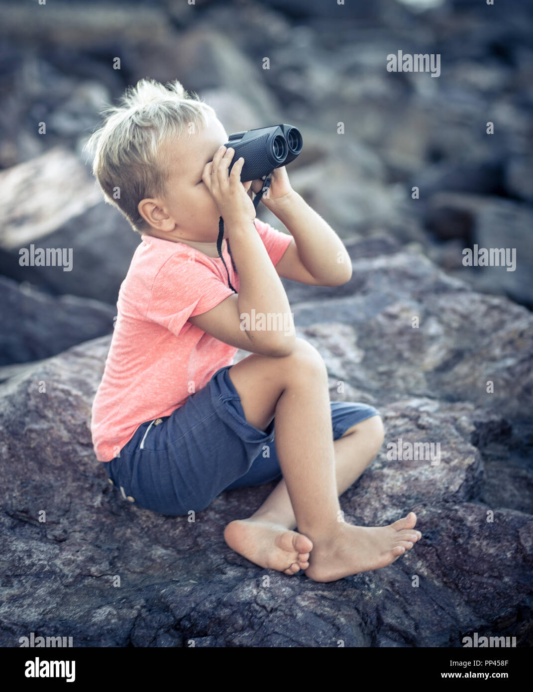 Chico busca lejos con binoculares, sentado sobre una roca cerca del mar Foto de stock