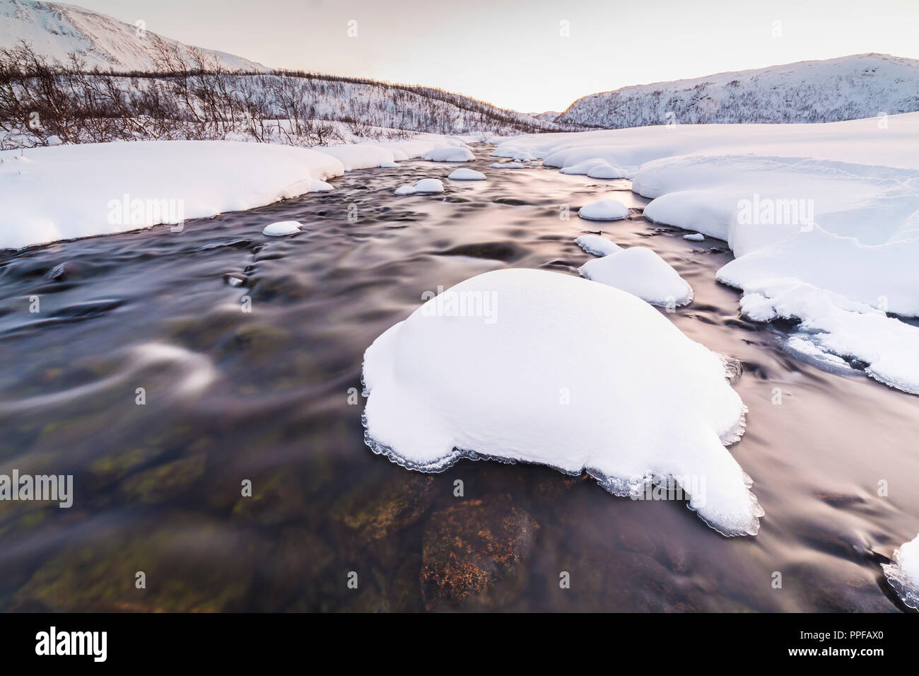 Un poco de agua corriente con rocas en primer plano con un paisaje nevado de invierno ártico, en el fondo. El cielo está despejado, múltiple y vibrante col Foto de stock
