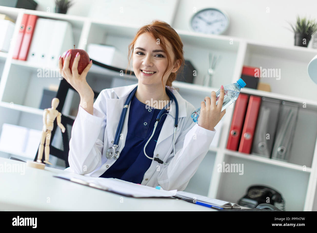 Una hermosa joven vestida con una túnica blanca se sienta en una mesa en la oficina y tiene una manzana y una botella de agua en su mano. Foto de stock
