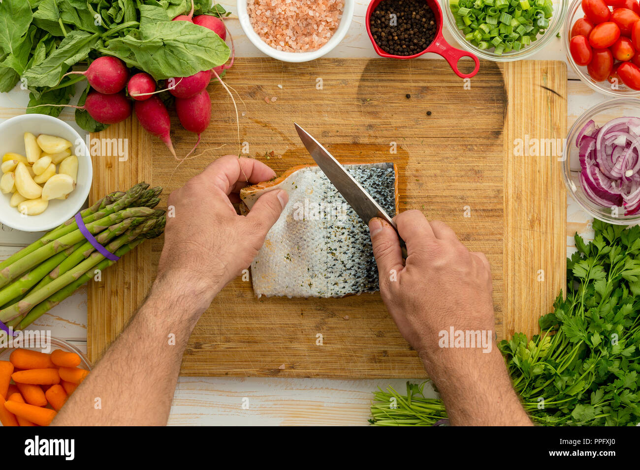 Hombre preparar un filete de salmón fresco para cocinar la limpieza de la piel con un cuchillo afilado en una placa rodeada por frescas ensaladas, cerrar overhea Foto de stock