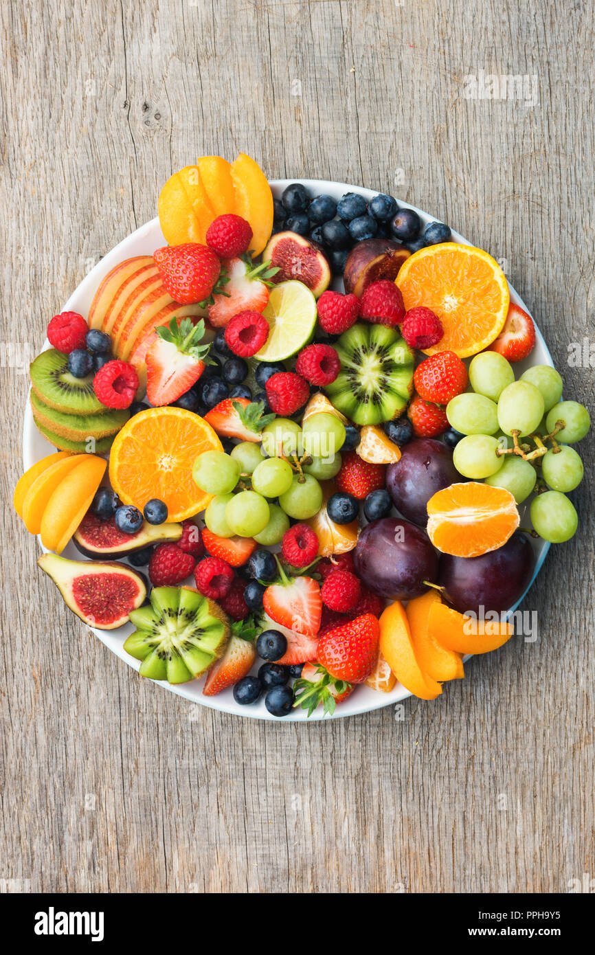 Plato de frutas saludables frambuesas, fresas, naranjas Ciruelas Kiwis Manzanas Uvas arándanos sobre la mesa de madera de color gris oscuro, vertical, vista superior, copia Foto de stock