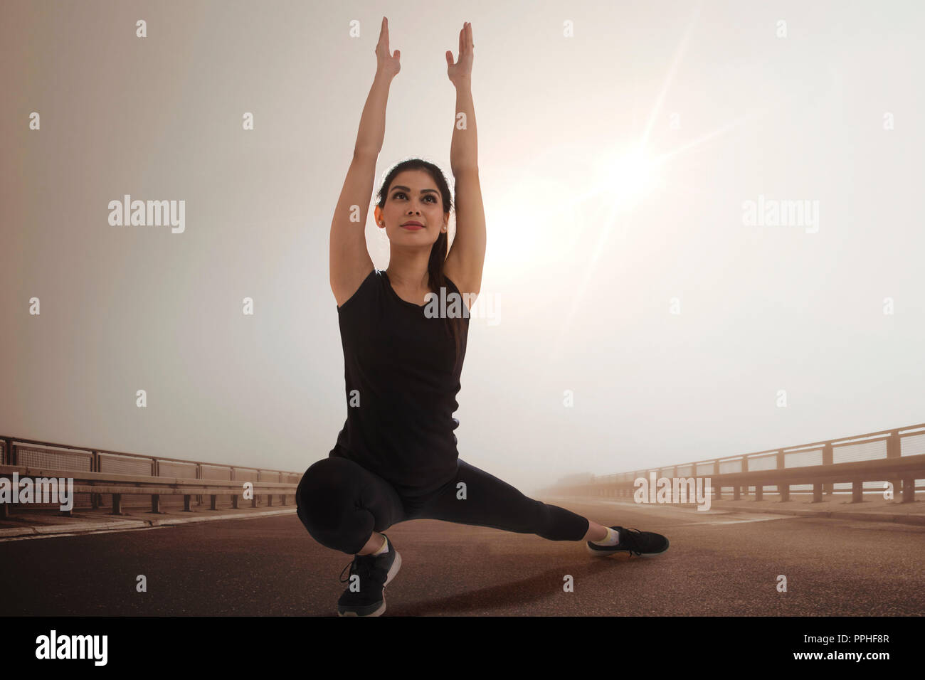 Mujer joven en ropa de entrenamiento haciendo ejercicios de estiramiento al aire libre con el sol en el fondo. Foto de stock