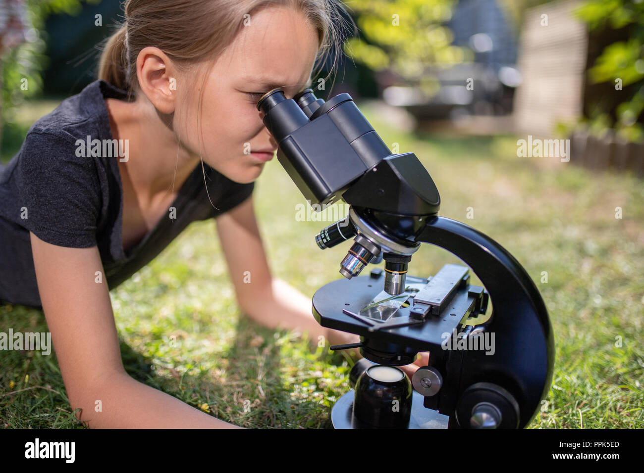Una niña de 9 años se encuentra en la hierba en el jardín y se ve en el ocular de un microscopio. Foto de stock