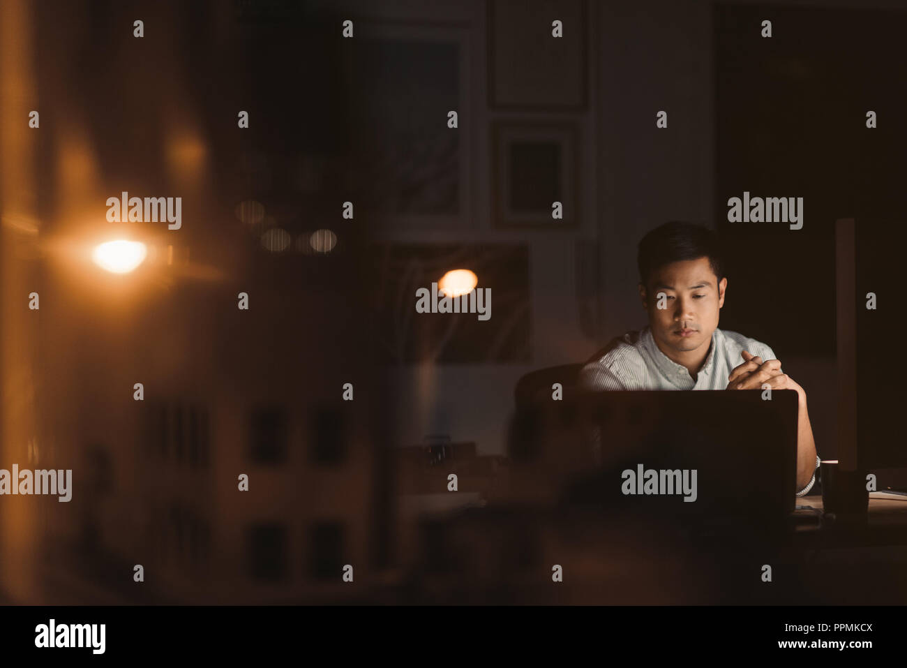 Joven empresario asiático sentado en su mesa de trabajo en un ordenador portátil tarde en la noche por las ventanas de las oficinas que refleja las luces de la ciudad Foto de stock