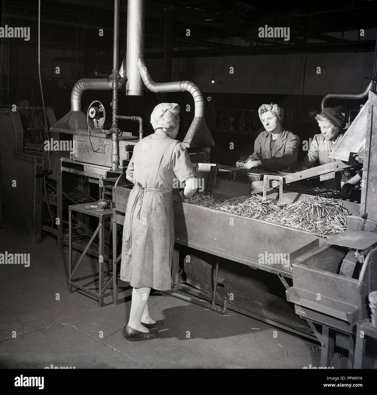 1950s, históricos, las mujeres que trabajan en las fábricas con un mono y  cabello permanente bufandas por bandejas de componentes metálicos  producidos por la maquinaria al fabricante de pilas de cinc-carbono, siempre