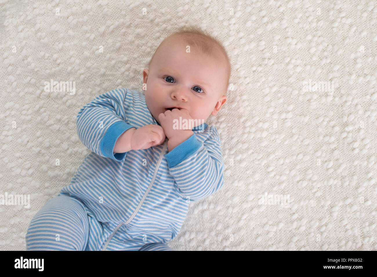 Un bebé de 2 meses niño acostado sobre la espalda en una sábana blanca. Él  está usando pijama de rayas azul y blanco y tiene su mano en su boca y está
