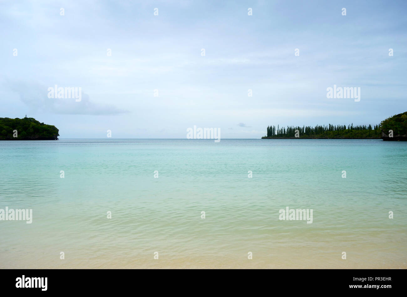 Aqua-claras aguas de color se extienden desde una playa para los cabeceros en la Isla de Pinos. Más allá está el Océano Pacífico Sur. El cielo es azul con suaves cl Foto de stock