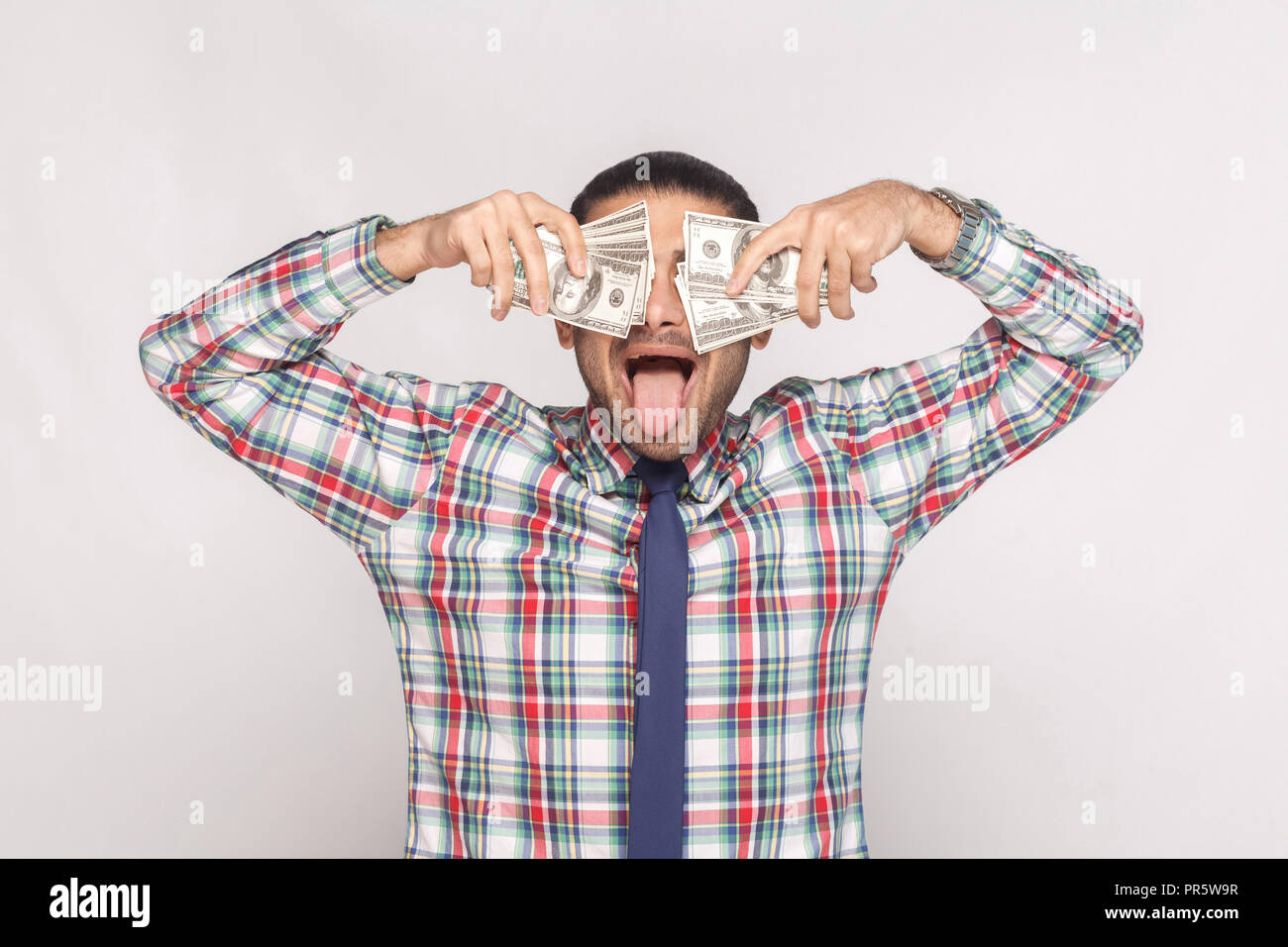 Enloquece con éxito! Ricos apuesto hombre barbado en coloridos camisa a cuadros con corbata azul permanente y tapar los ojos con ventilador de dinero con lengua ou Foto de stock