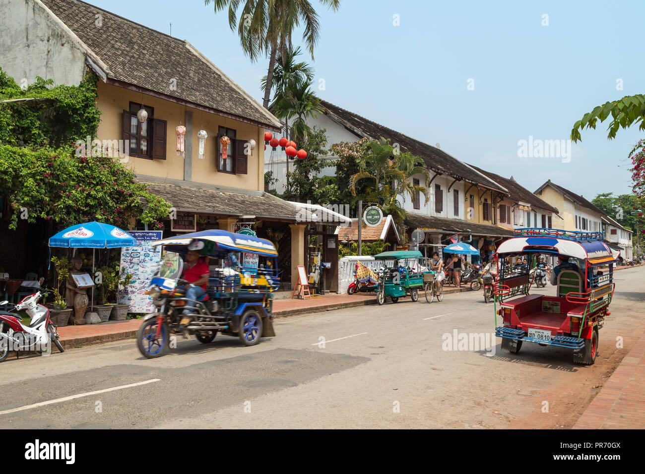 Bicicletas, motos y dos coloridos taxis de tres ruedas llamado jumbo (o un tuk-tuk) en la calle Sisavangvong en Luang Prabang, Laos, en un día soleado. Foto de stock