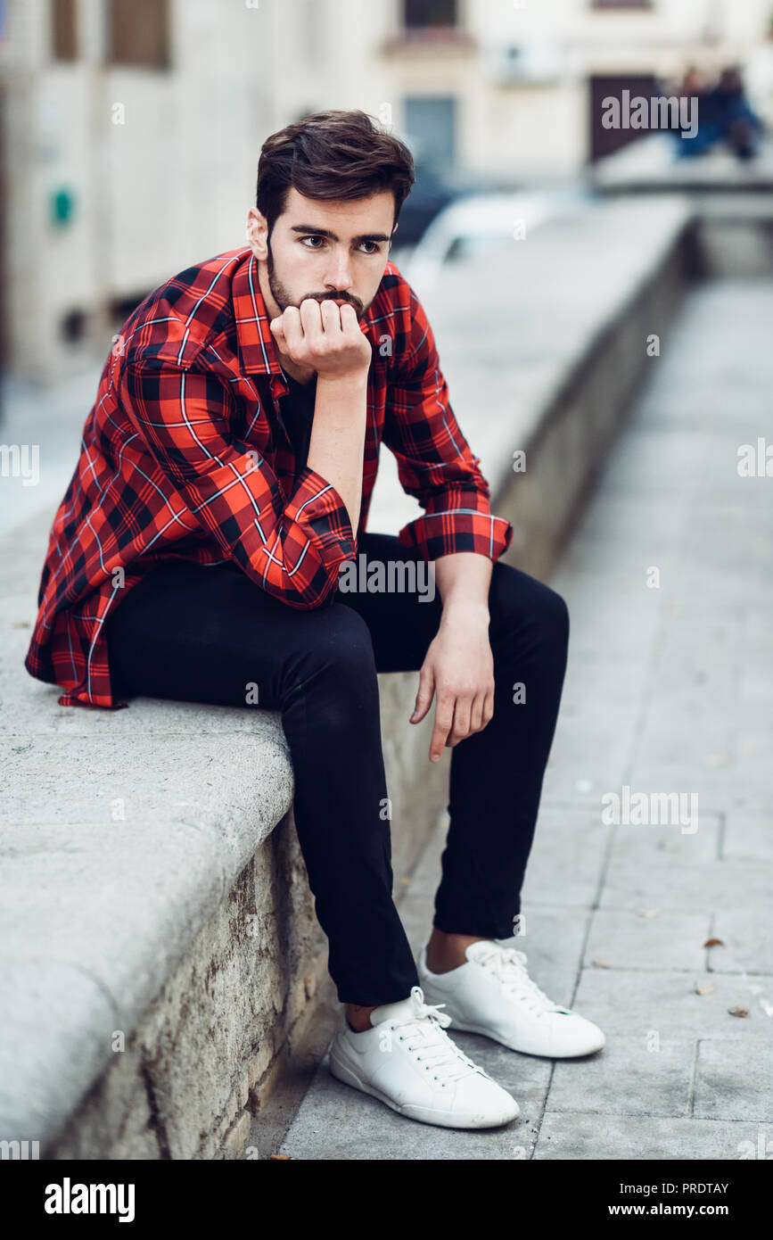 Joven Hombre Barbado, modelo de moda, sentado en el fondo urbano vistiendo  ropa casual. Chico con barba y peinado moderno en la calle con el Plaid  Fotografía de stock - Alamy