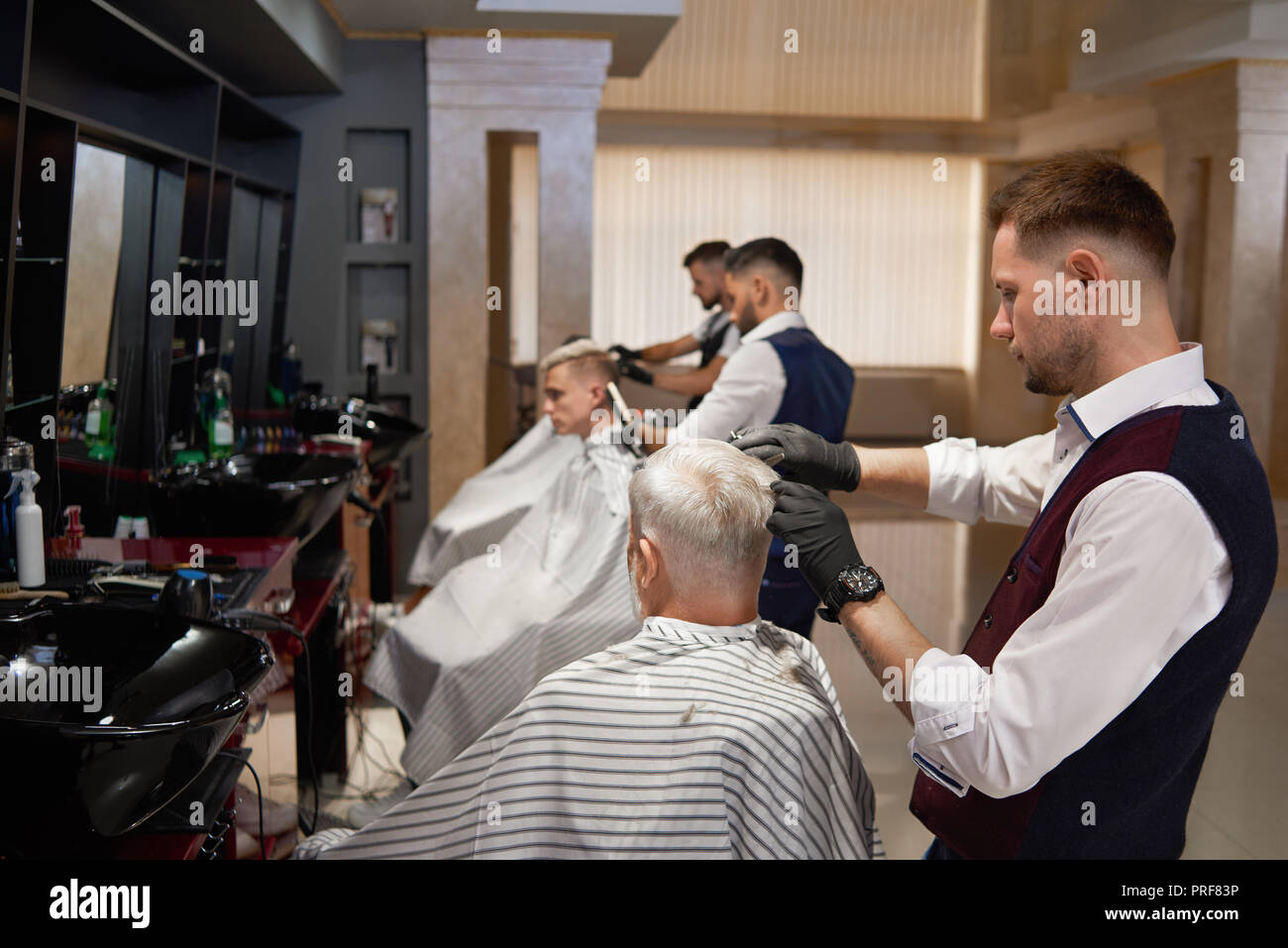 Vista lateral de la peluquería masculina profesional trabajando en el  barbershop moderno con los clientes. Máster en diseño uniforme y peinado  corte de pelo de los clientes masculinos que sentado en trajes