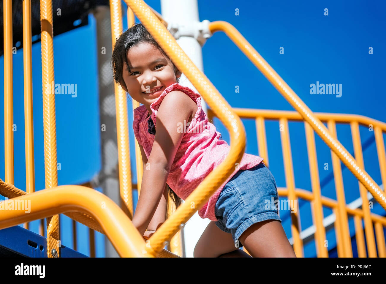 Linda chica subiendo un colorido parque infantil usando las manos y los pies con una sonrisa en su rostro, mostrando excelentes habilidades motoras Foto de stock