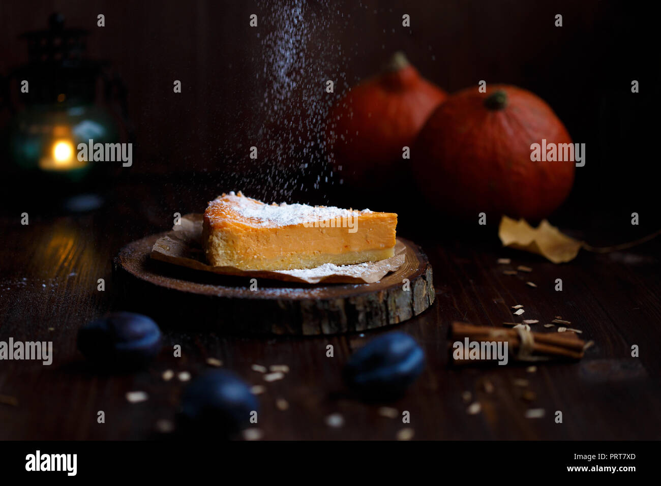 Lado femenino espolvorea azúcar en polvo sobre el cheesecake de calabaza. Calabazas, lámpara de mesa, follaje, madera de vainilla sobre un fondo oscuro. El otoño y el invierno cálido concepto Foto de stock