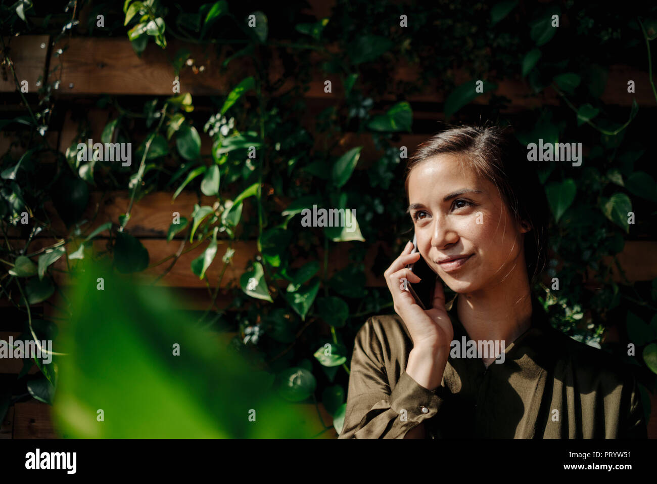 Mujer sonriente en celular en la pared con plantas trepadoras Foto de stock