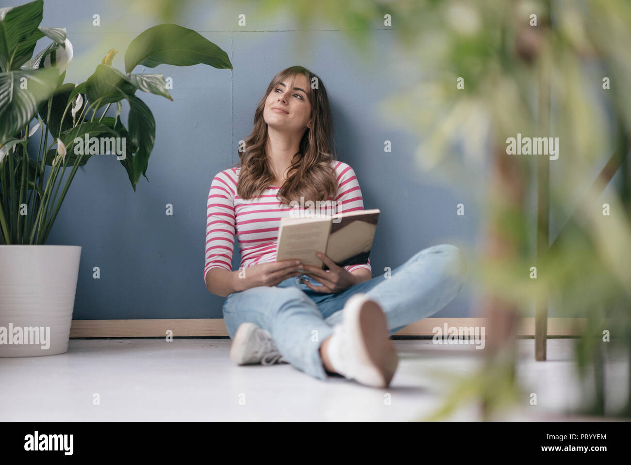 Mujer sentada en el suelo en su nueva casa, leer un libro, rodeado de plantas Foto de stock