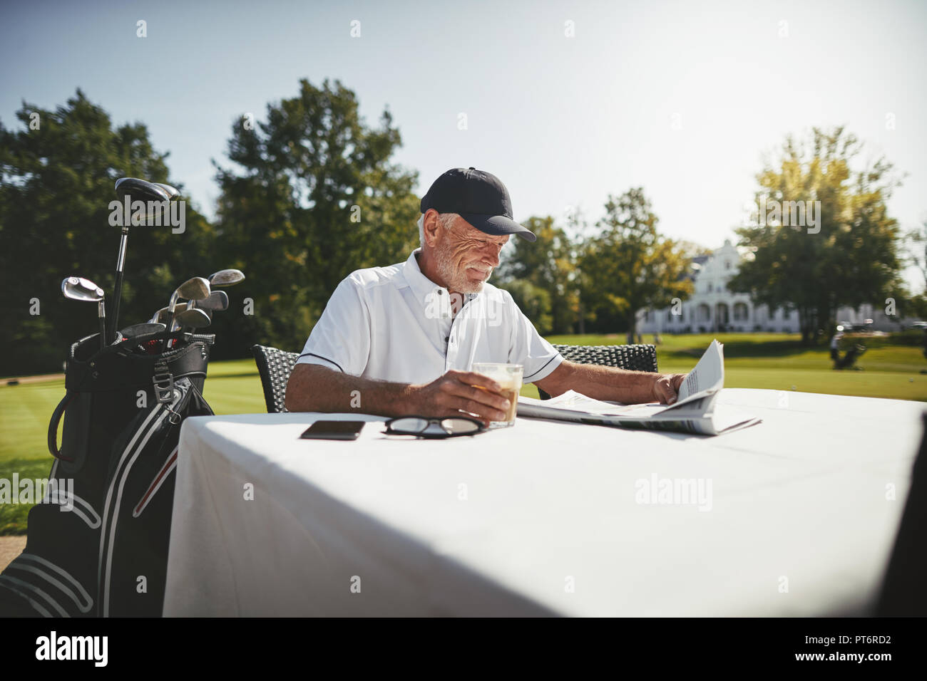 Senior sonriente hombre disfrutando de un café y leyendo un periódico mientras se relaja en un supuesto restaurante después de jugar una ronda de golf Foto de stock