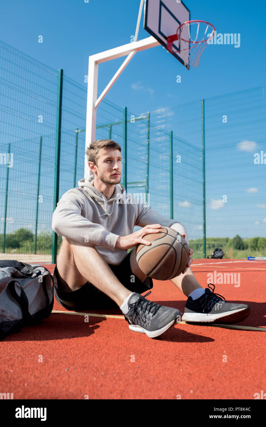 Longitud total retrato contemporáneo del hombre joven sentado en el suelo  en la cancha de baloncesto sosteniendo la bola, espacio de copia Fotografía  de stock - Alamy