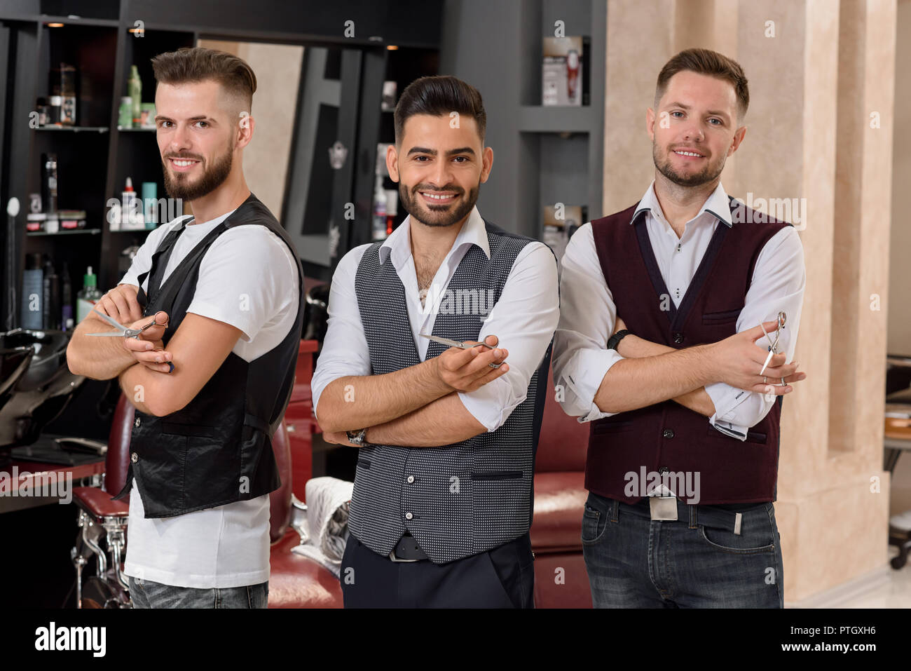 Barberos mostrando posando con manos cruzadas y mirando a la cámara. Guapo peluqueros vistiendo camisas blancas y chalecos. Maestría profesional y permanente posando en el barbershop. Foto de stock