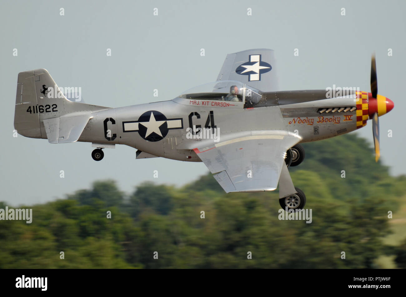 North American P-51 Mustang avión de combate llamado Nooky Booky IV volando en un festival aéreo. Pasado volando bajo los árboles. Abajo del tren de rodaje. Espacio para copiar Foto de stock