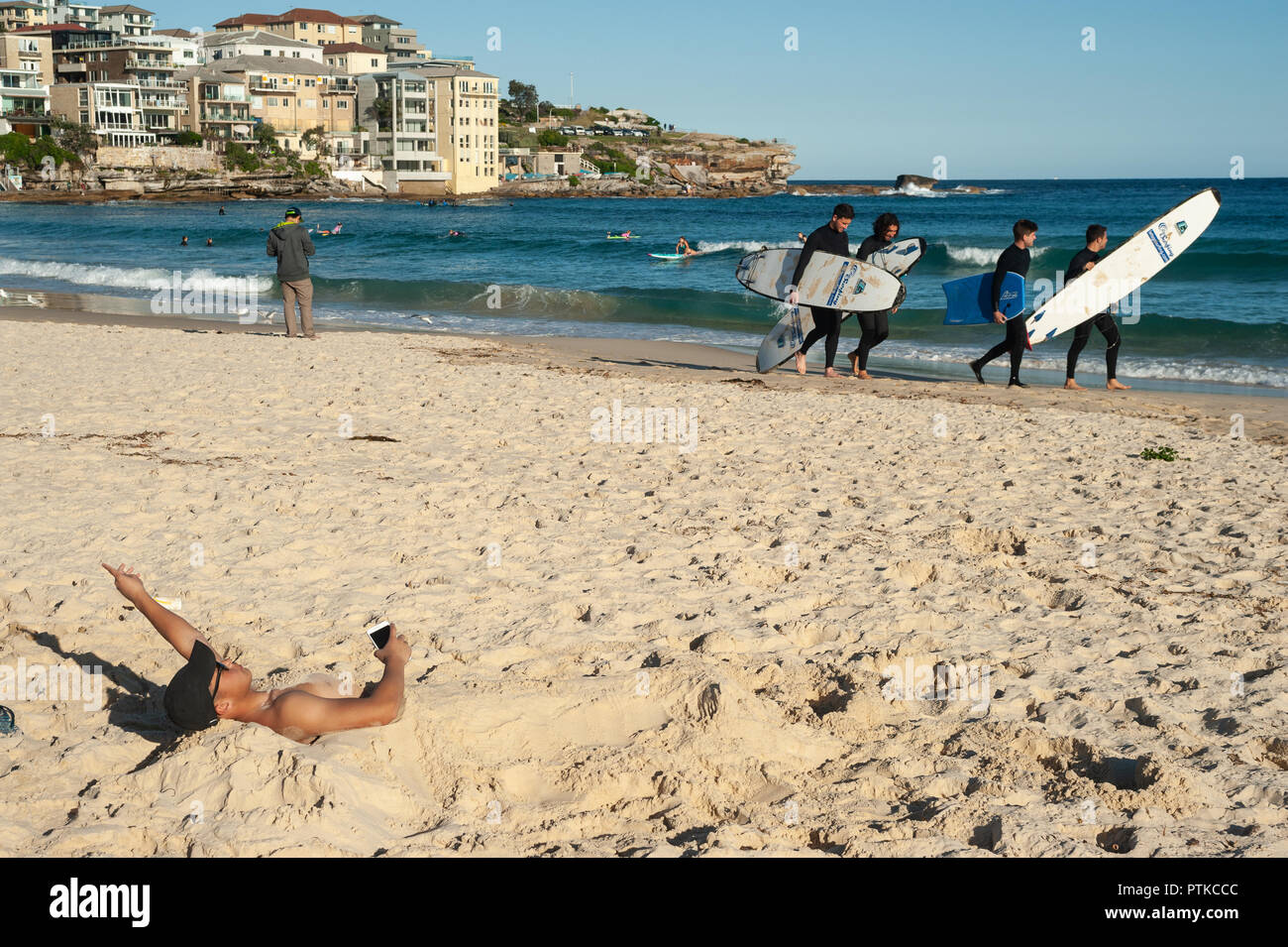 21.09.2018, Sydney, New South Wales, Australia - un joven se ve medio enterrado en la arena en la playa de Bondi, como un grupo de jóvenes surfistas camina por. Foto de stock