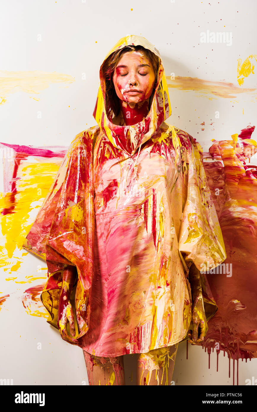 Hermosa mujer de impermeable pintadas con pintura amarilla y roja de pie con los ojos cerrados Foto de stock