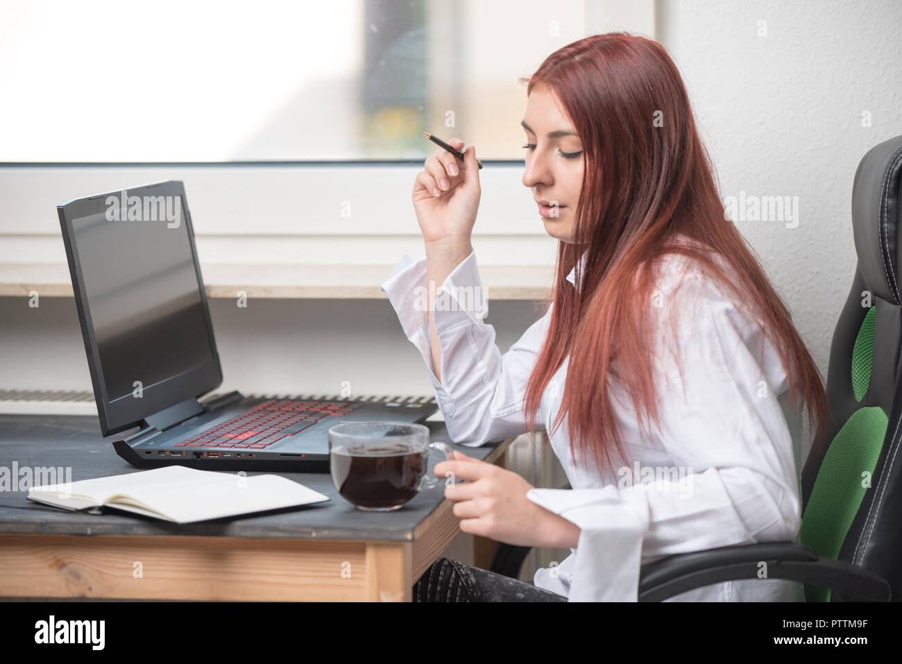 Un joven, atractivo, feliz mujer está sentado detrás de un escritorio por la ventana. Ella trabaja. Abrir un portátil, el café y el portátil en el escritorio Foto de stock