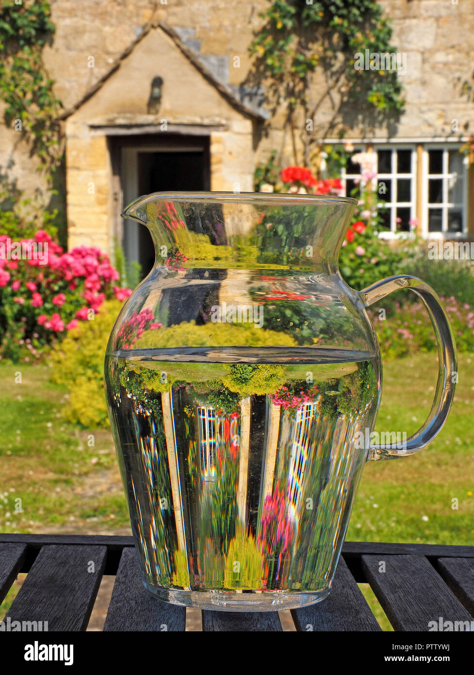 Jarra de agua de cristal refracte imagen de flores en inglés Country Garden  & color miel de casa en piedra con ventanas parteluz en Cotswolds,  Inglaterra, Reino Unido Fotografía de stock -