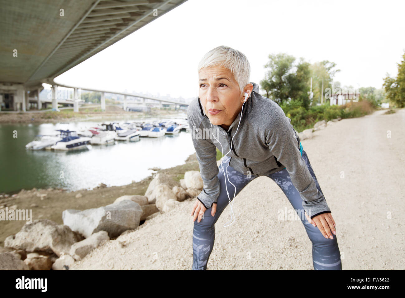 Activa ejecutando Senior Mujer descansando después de correr escuchando música con el smartphone en la orilla del río , entrenar al aire libre. Foto de stock